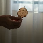 Immagine simbolica della fragilità in un paziente anziano con BPCO: una mano anziana che tiene delicatamente una foglia secca e fragile, illuminata da una luce naturale morbida che entra da una finestra. Obiettivo macro 100mm, alta definizione, messa a fuoco selettiva sulla mano e sulla foglia per enfatizzare la delicatezza.