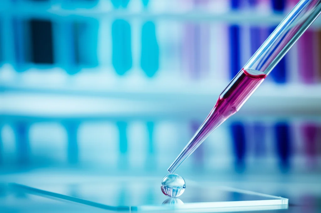 Fotografia macro di una goccia di sangue su un vetrino da microscopio, con pipette e provette sfocate sullo sfondo in un laboratorio. Lente macro 90mm, illuminazione controllata, alta definizione per evidenziare i dettagli del sangue.