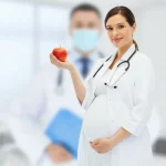 Donna incinta sorridente che tiene in mano una mela rossa, simbolo di salute, con alle spalle uno sfondo medico astratto e leggermente sfocato sui toni del blu e del bianco, obiettivo 50mm, luce naturale morbida laterale, colori caldi e accoglienti, profondità di campo media.