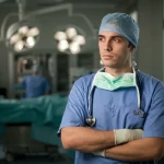 Immagine fotorealistica di uno specializzando in chirurgia pensieroso in un corridoio d'ospedale, luce soffusa proveniente da una finestra, sullo sfondo sfocato si intravede una sala operatoria. Obiettivo 50mm, profondità di campo, toni leggermente freddi per suggerire stress ma anche riflessione.