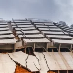 Immagine fotorealistica del sito di un parcheggio multipiano crollato su un pendio montuoso in India, vista grandangolare 10mm che mostra l'entità del danno, il muro di contenimento in cemento armato rotto, il terreno franato e le strutture danneggiate, sotto un cielo nuvoloso post-pioggia. Long exposure per nuvole mosse, sharp focus sul disastro.