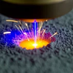Primo piano macro di un processo di fusione laser su letto di polvere (PBF-LB), il raggio laser blu intenso colpisce la polvere metallica grigia scura creando una pozza fusa incandescente di colore giallo-arancio con scintille luminose che si sollevano. Obiettivo macro 100mm, alta definizione, illuminazione controllata laterale per evidenziare la texture della polvere e la fluidità del metallo fuso, sfondo sfocato per concentrare l'attenzione sull'interazione laser-materiale.
