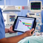 Immagine fotorealistica di un medico in terapia intensiva che consulta un tablet mostrando un nomogramma di dosaggio per il meropenem, accanto a un letto di paziente collegato a una macchina SLEDD. Obiettivo prime 35mm, profondità di campo che sfoca leggermente lo sfondo dell'ICU, illuminazione mista clinica e dello schermo.