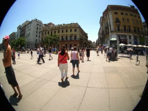 Veduta grandangolare di un paesaggio urbano durante un'ondata di calore estiva opprimente. Aria tremolante per il caldo, persone che cercano ombra, cielo terso e sole accecante. Sensazione di disagio e calore intenso. Obiettivo grandangolare 20mm, messa a fuoco nitida sull'intera scena.