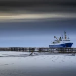 Ampia veduta del Mare di Barents al tramonto o all'alba, con un peschereccio da granchi delle nevi in lontananza che cala o recupera le sue lunghe file di nasse. Wide-angle 10-24mm, lunga esposizione per rendere l'acqua liscia e il cielo suggestivo, focus nitido sulla scena, luce drammatica che evoca la vastità e le sfide dell'ambiente.