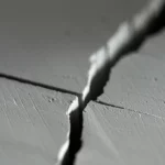 Immagine macro fotorealistica di un materiale piezoelettrico ceramico (tipo PZT) con una crepa rettangolare ben definita al centro. Illuminazione laterale radente per esaltare la texture e la tridimensionalità della crepa. Obiettivo macro 100mm, alta definizione, messa a fuoco selettiva sulla punta della crepa, sfondo leggermente sfocato. Dettagli microscopici visibili.