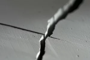 Immagine macro fotorealistica di un materiale piezoelettrico ceramico (tipo PZT) con una crepa rettangolare ben definita al centro. Illuminazione laterale radente per esaltare la texture e la tridimensionalità della crepa. Obiettivo macro 100mm, alta definizione, messa a fuoco selettiva sulla punta della crepa, sfondo leggermente sfocato. Dettagli microscopici visibili.