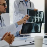 Un gruppo eterogeneo di medici esperti in una sala riunioni luminosa, intenti a esaminare radiografie e dati su un tablet, discutendo una diagnosi complessa. Obiettivo prime 35mm, luce naturale, profondità di campo.