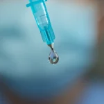 Fotografia macro di una goccia di lidocaina che cade dalla punta di una siringa, con sfondo sfocato di un ambiente medico sterile. Illuminazione drammatica e controllata, alta definizione, focus estremamente preciso sulla goccia. Lente macro 105mm.