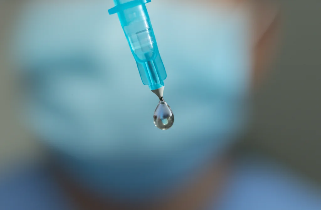 Fotografia macro di una goccia di lidocaina che cade dalla punta di una siringa, con sfondo sfocato di un ambiente medico sterile. Illuminazione drammatica e controllata, alta definizione, focus estremamente preciso sulla goccia. Lente macro 105mm.