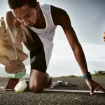 Fotografia sportiva di un velocista sulla linea di partenza accanto a un maratoneta in corsa su una strada panoramica, immagine divisa o composita, luce naturale drammatica, teleobiettivo zoom 150mm, fast shutter speed per catturare il movimento del maratoneta e la tensione del velocista.