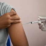 Primo piano di un braccio di un adulto indiano che riceve una vaccinazione da un operatore sanitario in una clinica a Nuova Delhi. Obiettivo prime 35mm, profondità di campo per mettere a fuoco l'iniezione, luce naturale soffusa.