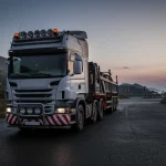 Immagine fotorealistica di un moderno camion articolato a cinque assi con tre assi sterzanti che manovra con precisione su una strada tortuosa in montagna al crepuscolo. Obiettivo prime 35mm, profondità di campo che sfoca leggermente lo sfondo montano, illuminazione drammatica con fari accesi, dettagli elevati sul veicolo e sulla texture della strada bagnata.