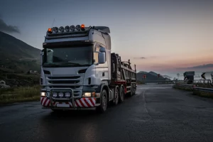 Immagine fotorealistica di un moderno camion articolato a cinque assi con tre assi sterzanti che manovra con precisione su una strada tortuosa in montagna al crepuscolo. Obiettivo prime 35mm, profondità di campo che sfoca leggermente lo sfondo montano, illuminazione drammatica con fari accesi, dettagli elevati sul veicolo e sulla texture della strada bagnata.