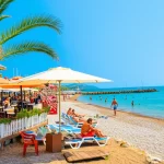 Veduta panoramica di una vivace località costiera turca sotto il sole estivo, con turisti che si rilassano in aree ombreggiate vicino a caffè all'aperto e altri che passeggiano vicino al mare cristallino sulla spiaggia sabbiosa. Fotografia di paesaggio, obiettivo grandangolare 15mm, messa a fuoco nitida, colori vividi e brillanti tipici del Mediterraneo.