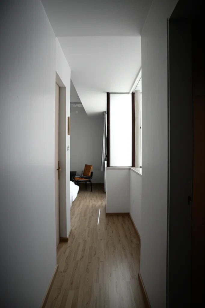 Fotografia architettonica di un interno modernista con focus sulla divisione degli spazi, obiettivo 35mm, luce soffusa che entra da una finestra laterale, creando un gioco di luci e ombre che enfatizza la separazione tra una zona giorno e un corridoio che porta alla zona notte, evocando il concetto di privacy visiva analizzata tramite Space Syntax in edifici residenziali.