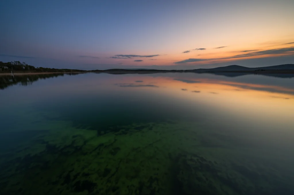 Fotografia paesaggistica grandangolare di un lago meromictico al tramonto, con acqua calma che riflette il cielo, obiettivo 15mm, lunga esposizione per acqua liscia, focus nitido, che evoca gli oceani primordiali anossici.