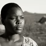 Immagine fotorealistica di una donna sudafricana in un contesto rurale, il suo volto esprime forza mista a preoccupazione, luce naturale filtrata del tardo pomeriggio che crea lunghe ombre. Obiettivo da ritratto 35mm, leggero effetto duotone seppia e grigio per dare un tono riflessivo, profondità di campo che sfoca lo sfondo di un paesaggio arido con capanne tradizionali.
