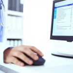 Immagine fotorealistica, obiettivo prime 35mm con profondità di campo, che mostra la mano di un medico che utilizza un mouse su un computer. Sullo schermo è visibile un software di prescrizione elettronica con un elenco di esami di laboratorio e i relativi prezzi evidenziati accanto a ciascuna voce. L'ambiente è un ufficio medico moderno e luminoso. Toni di colore neutri con accenti blu sullo schermo.