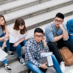 Fotografia stile ritratto di gruppo, leggermente dall'alto, di giovani studenti universitari asiatici seduti sui gradini, con espressioni miste che suggeriscono dinamiche sociali e preoccupazioni sull'immagine corporea. Obiettivo 35mm, luce naturale, stile fotorealistico.