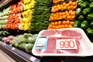 Immagine fotorealistica, lente primaria 50mm, attenzione selettiva su un prezzo di prezzo del supermercato collegato a un pacchetto di carne rossa, il prezzo leggermente elevato. In background sfocato, gli scaffali forniti di frutta e verdura colorate sono visibili, simboleggiano la scelta tra carne e opzioni a base vegetale e l'impatto delle potenziali tasse sulla carne o la rimozione dei sussidi. Illuminazione controllata e neutra del supermercato.