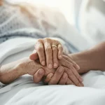 Mani di diverse tonalità di pelle che si stringono delicatamente la mano di un paziente anziano a letto in un ambiente ospedaliero luminoso, obiettivo 50mm, profondità di campo per mettere a fuoco le mani ma sfocare leggermente lo sfondo, simboleggiando cura, diversità ed empatia nelle cure palliative.