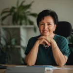 Ritratto fotografico di una donna sorridente di circa 58 anni, elegantemente vestita, seduta alla sua scrivania in un ufficio con piante sullo sfondo. Guarda verso la camera con espressione fiduciosa e serena. Obiettivo prime 50mm, luce morbida laterale, profondità di campo che sfoca leggermente lo sfondo, toni caldi.