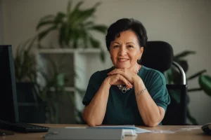 Ritratto fotografico di una donna sorridente di circa 58 anni, elegantemente vestita, seduta alla sua scrivania in un ufficio con piante sullo sfondo. Guarda verso la camera con espressione fiduciosa e serena. Obiettivo prime 50mm, luce morbida laterale, profondità di campo che sfoca leggermente lo sfondo, toni caldi.