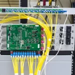Fotografia dettagliata del prototipo dello switch core-selettivo all-fiber montato su un rack standard da 19 pollici in un ambiente di laboratorio. Si intravedono le connessioni in fibra ottica gialla, i moduli dei modulatori di fase e i componenti elettronici di controllo su una scheda. Obiettivo prime 50mm, illuminazione da studio controllata, focus preciso sul dispositivo assemblato.