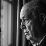 Ritratto fotografico di un uomo anziano (circa 80 anni) dall'aspetto riflessivo, visto di profilo vicino a una finestra con luce morbida che ne illumina parzialmente il volto. Obiettivo prime 35mm, bianco e nero film, profondità di campo che sfoca leggermente lo sfondo esterno alla finestra.