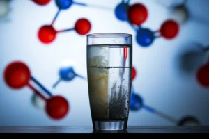 Fotografia concettuale wide-angle che mostra un bicchiere d'acqua limpida per metà e torbida per l'altra metà, simboleggiando il processo di purificazione. Sullo sfondo, una sfocata rappresentazione di molecole di Sulfametossazolo e particelle di Ag3PO4 interagenti sotto una luce stilizzata. Obiettivo 20mm, lunga esposizione per un effetto etereo sull'acqua, focus nitido sulla linea di separazione tra acqua pulita e contaminata.