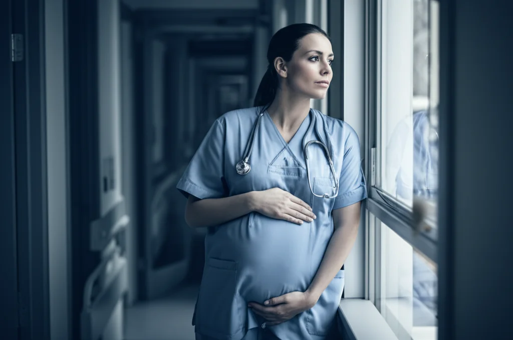Fotografia stile ritratto di un'infermiera incinta che guarda pensierosa fuori da una finestra d'ospedale durante una pausa. Obiettivo 35mm, toni duotone blu e grigio, profondità di campo che sfoca l'esterno.