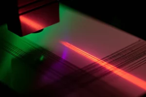 Immagine fotorealistica di un raggio laser (mostrato come un fascio di luce astratta che sfuma tra UV-viola, Visibile-verde e IR-rosso) che colpisce la superficie di un campione metallico lucido (lega Fe-20Mn), creando micro-scanalature brillanti e precise. Stile still life high-tech, obiettivo macro 85mm, illuminazione drammatica controllata che evidenzia la texture, alta definizione dei dettagli sulla superficie metallica e sull'interazione laser.