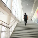 Una persona vista di spalle che sale con passo deciso una luminosa rampa di scale in un edificio moderno. Accanto alle scale, un grande banner colorato con messaggi motivazionali sull'attività fisica è chiaramente visibile. Obiettivo prime 35mm, luce naturale diffusa, profondità di campo che mantiene a fuoco sia la persona che il banner.