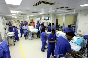 Fotografia grandangolare, obiettivo 18mm, che mostra l'interno di un pronto soccorso affollato di un ospedale safety-net. Si vedono persone di diverse etnie, sia pazienti su barelle che personale medico indaffarato. L'illuminazione è quella tipica ospedaliera, un po' fredda. L'immagine trasmette un senso di urgenza, complessità e diversità. Messa a fuoco nitida su tutta la scena per catturare i dettagli dell'ambiente.