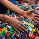 Immagine fotorealistica di mani esperte che smistano meticolosamente scarti tessili colorati in un centro di riciclaggio indiano ben illuminato. Dettaglio elevato sui tessuti, obiettivo macro 85mm, luce controllata che evidenzia le texture diverse e simboleggia lo sforzo verso l'economia circolare.