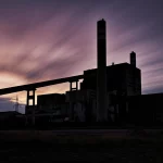 Fotografia paesaggistica, obiettivo grandangolare 18mm, di un'imponente centrale elettrica a combustibili fossili dismessa al crepuscolo, silhouette scura contro un cielo colorato, lunga esposizione per nuvole fluide, focus nitido sull'architettura industriale decadente, atmosfera malinconica.