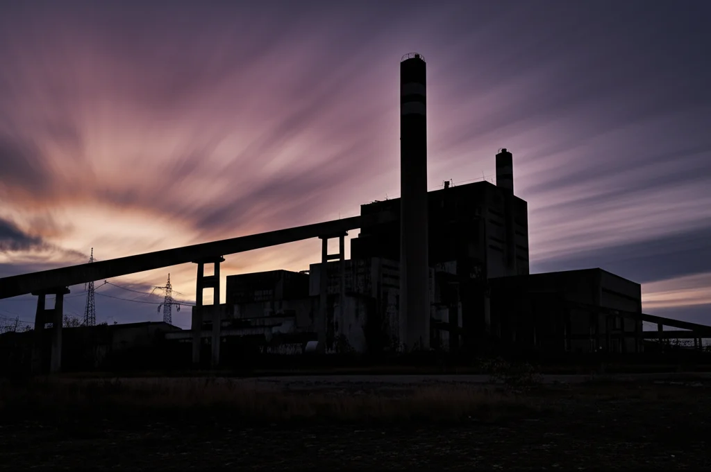 Fotografia paesaggistica, obiettivo grandangolare 18mm, di un'imponente centrale elettrica a combustibili fossili dismessa al crepuscolo, silhouette scura contro un cielo colorato, lunga esposizione per nuvole fluide, focus nitido sull'architettura industriale decadente, atmosfera malinconica.