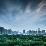 Skyline di una metropoli cinese moderna al tramonto, con ciminiere industriali in lontananza parzialmente oscurate da parchi urbani verdi in primo piano, simboleggiando la sfida e l'obiettivo della trasformazione verso città a rifiuti zero. Obiettivo grandangolare 20mm, long exposure per nuvole soffici, focus nitido sull'intera scena.