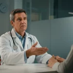 Farmacista in camice bianco che consulta un paziente in un ambiente clinico luminoso e moderno, discutendo di farmaci. Obiettivo prime 50mm, luce naturale laterale, focus sul dialogo e l'interazione.