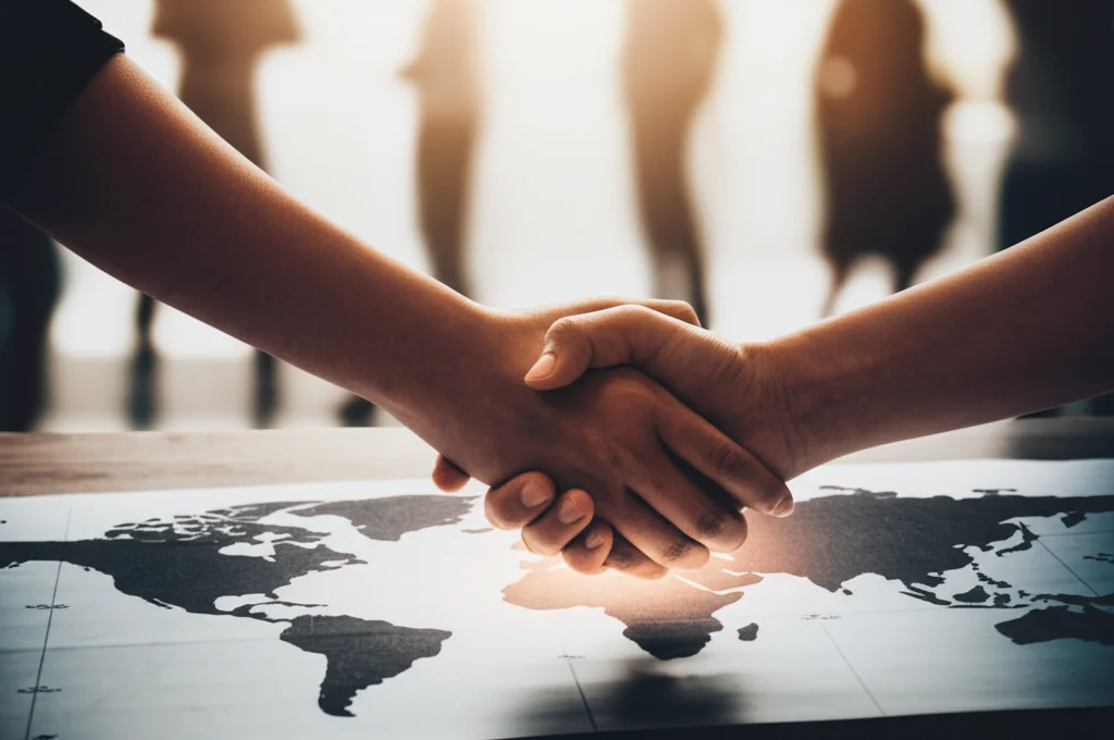 Fotografia concettuale, obiettivo prime 50mm con profondità di campo media. Due mani di etnie diverse si stringono sopra una mappa stilizzata del mondo, illuminata da una luce calda che simboleggia la speranza e la solidarietà. Sullo sfondo, sagome sfocate di persone in movimento. Toni caldi e leggermente desaturati.