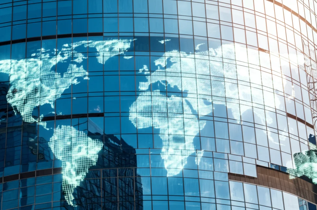 Immagine fotorealistica di un grattacielo moderno con riflessi di mappe globali sulle finestre, simboleggia l'espansione internazionale delle aziende e la comunicazione con i mercati finanziari. Obiettivo zoom 24mm, luce del tardo pomeriggio, profondità di campo.