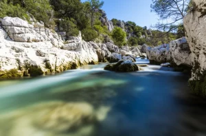 Fiume che scorre attraverso un paesaggio carsico con rocce calcaree bianche evidenti sulle sponde, obiettivo grandangolare 20mm, acqua limpida che riflette il cielo azzurro, luce solare brillante, messa a fuoco nitida su tutto il paesaggio, esposizione lunga per effetto seta sull'acqua.