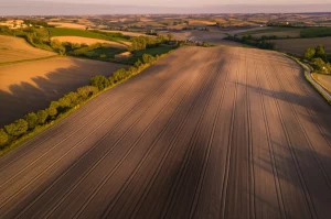 Immagine aerea ad alta risoluzione di un paesaggio italiano con campi coltivati e piccole strade, obiettivo grandangolare 15mm, lunga esposizione per nuvole morbide, si notano sottili linee parallele e perpendicolari nel terreno, indicative di antica centuriazione romana, luce dorata dell'alba.