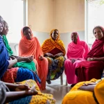 Un gruppo di donne etiopi sedute in cerchio in un ambiente comunitario semplice ma accogliente, che partecipano attivamente a una discussione di gruppo di supporto per il cancro al seno. Luce naturale morbida, espressioni di empatia e sostegno reciproco. Fotografia di ritratto di gruppo, obiettivo 35mm, profondità di campo per focalizzare sulle interazioni.
