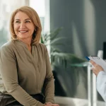 Una donna sopravvissuta al cancro al seno sorride con fiducia durante un controllo medico, luce naturale morbida che entra da una finestra, obiettivo prime 50mm, sfondo leggermente sfocato di uno studio medico accogliente e moderno, colori caldi e rassicuranti.