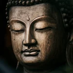 Immagine fotorealistica ad altissima definizione del volto enigmatico di una statua di Buddha asiatica antica, realizzata in pietra scura leggermente erosa dal tempo. Luce laterale drammatica che ne scolpisce i lineamenti sereni, obiettivo macro 100mm, messa a fuoco precisa sui dettagli degli occhi e delle labbra, sfondo scuro e sfocato.
