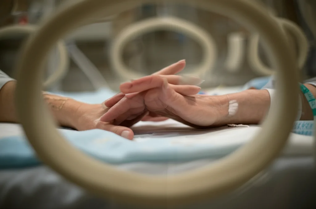 Immagine simbolica e delicata: le mani di una madre sfiorano delicatamente un'incubatrice in Terapia Intensiva Neonatale. Luce calda e soffusa, obiettivo macro 60mm, focus preciso sulle mani e sulla texture dell'incubatrice, sfondo sfocato per creare intimità e sottolineare il legame e la preoccupazione materna.