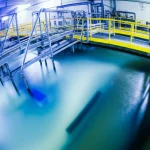 Vista grandangolare di un moderno impianto di trattamento delle acque reflue industriali, focalizzato su una vasca di reazione illuminata da luce UV, dove l'acqua bluastra viene trattata con elettrodi compositi titanio/grafite visibili. L'acqua nelle vasche successive appare progressivamente più chiara. Ambiente pulito e tecnologico, wide-angle 15mm, long exposure per acqua liscia, sharp focus sull'area di reazione.