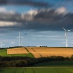 Fotografia paesaggistica con obiettivo grandangolare 20mm, che mostra un dilemma visivo: un bellissimo paesaggio naturale collinare tedesco parzialmente interrotto da moderne turbine eoliche in lontananza, luce drammatica all'alba, messa a fuoco nitida su tutto il campo, nuvole lisce da lunga esposizione, simboleggiando il trade-off tra energia rinnovabile e impatto visivo.